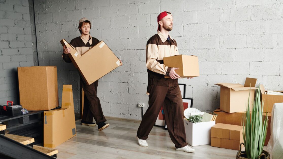 Two professional movers from a Moving service in New Jersey are carrying cardboard boxes inside a modern home with neatly stacked packing materials and organized supplies, indicating a smooth and efficient relocation process.