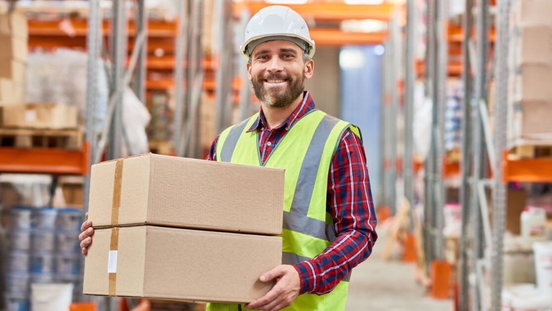 A smiling warehouse worker in a hard hat and safety vest holds two cardboard boxes inside a storage facility, representing the reliable and professional service of Jersey City movers.