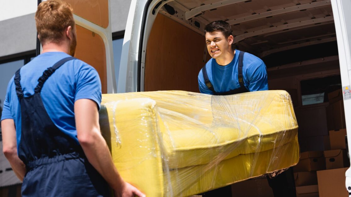 Two professional movers and packers in NJ wearing blue uniforms are carefully loading a yellow, plastic-wrapped couch into a moving van on a sunny day, showcasing efficient and reliable relocation services.