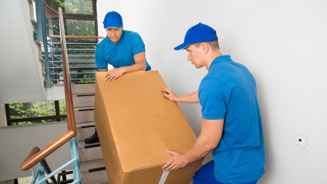 Two uniformed Movers NJ professionals in blue shirts and caps carefully carry a large, taped‑shut cardboard box up a bright residential staircase inside a building with windows.